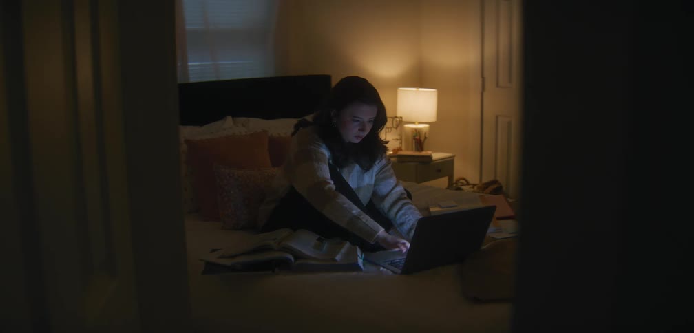 A girl sitting on a bed illuminated by the glow of a laptop screen as she studies