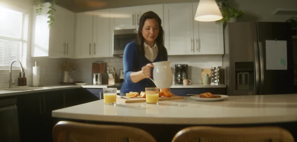 Woman standing in a kitchen pouring juice into a glass cup and slicing oranges on a countertop