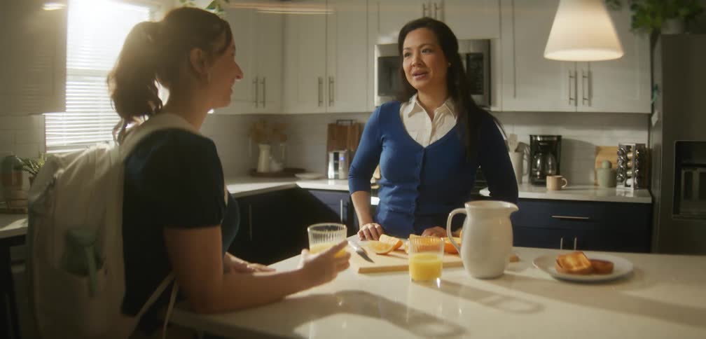 Young woman drinks orange juice while her mother leaves the kitchen