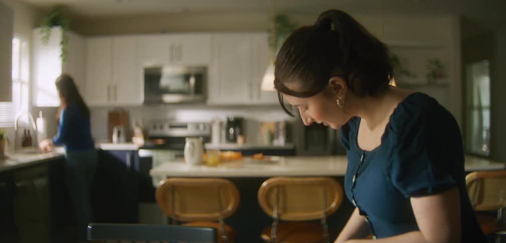 Mother preparing breakfast in the kitchen with her daughter placing her backpack on the chair at the table next to her