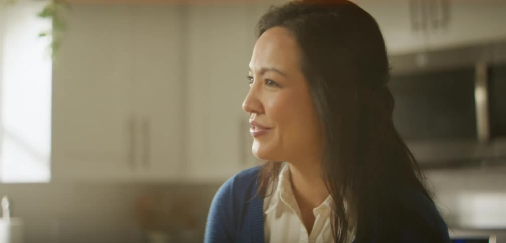 A woman is standing in her kitchen and talking to someone