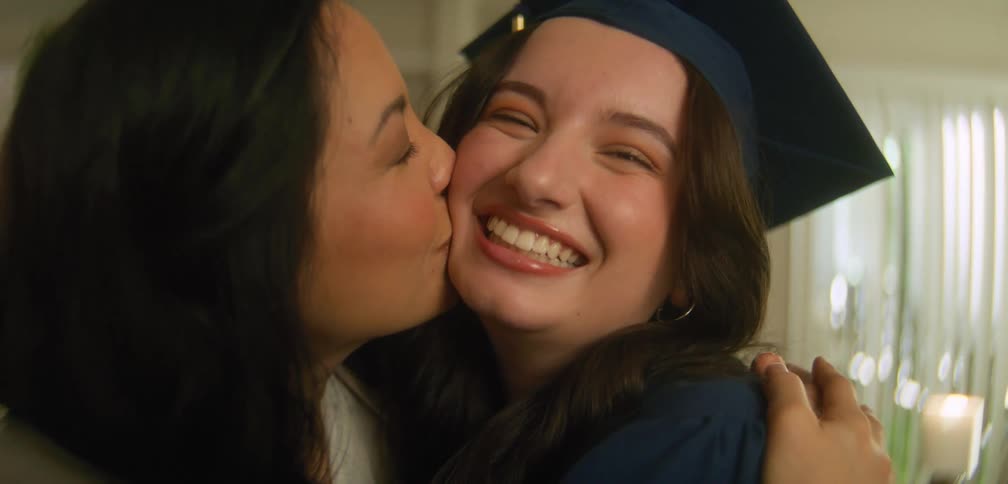Mother hugs and kisses her graduate daughter while they stand in their home