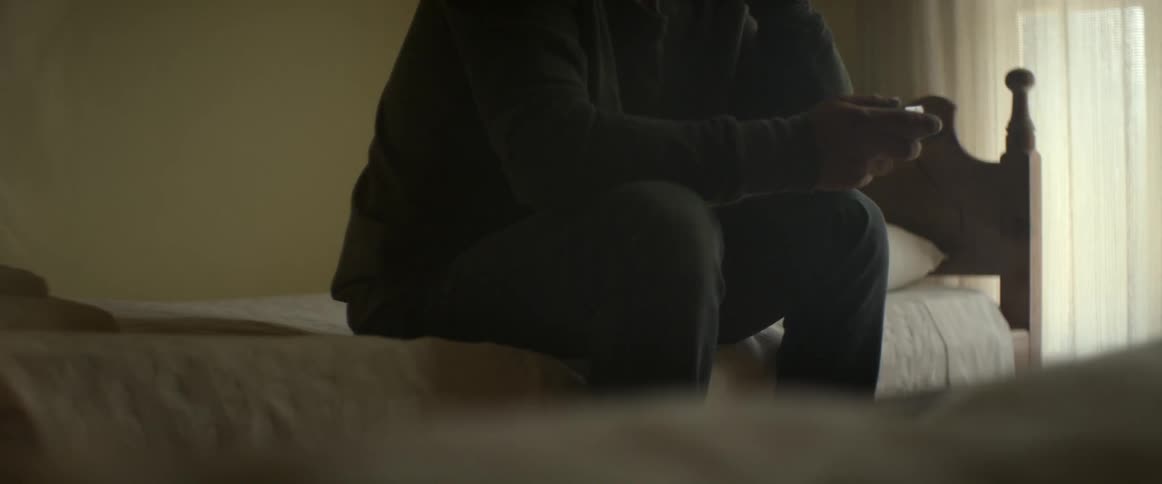 A man sitting nervously on the edge of a twin bed, holding a pack of cigarettes