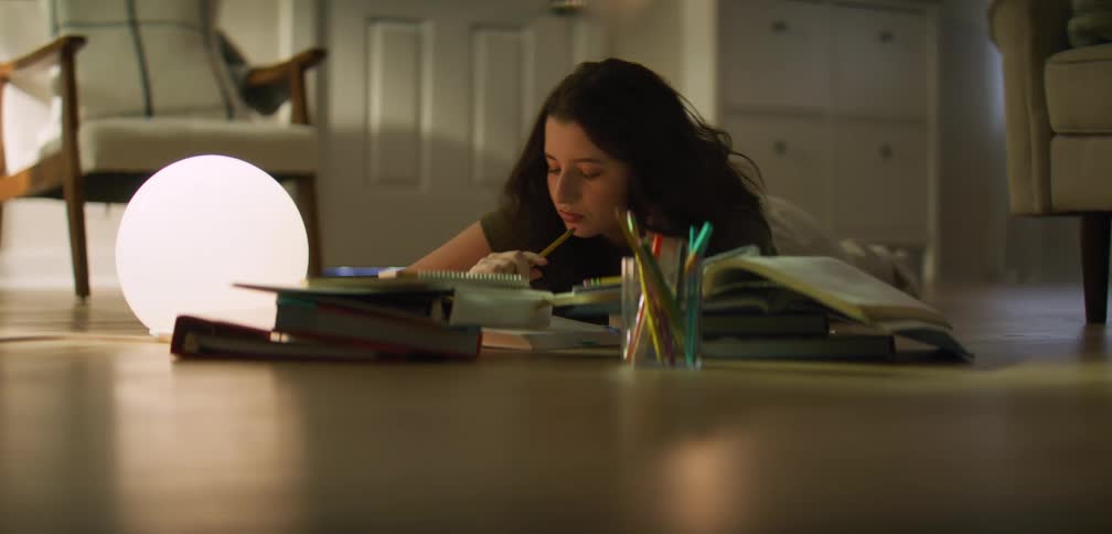 A young girl lying on the floor of a room with several books in front of her and she is studying