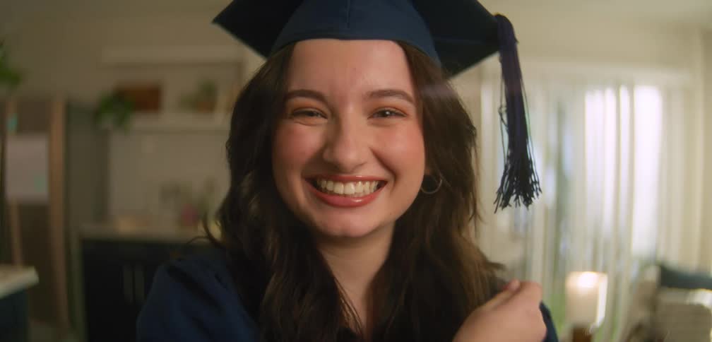 Female graduate standing in living room smiles while moving the tassel on her graduation cap