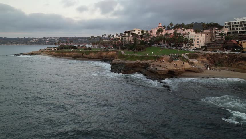 A rocky coastal shoreline with a green park area and pathways on the cliffside overlooking the ocean
