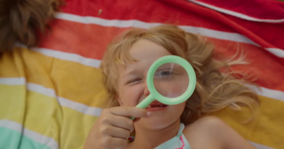 A girl with a magnifying glass over her eye lying on a colorful stripped blanket
