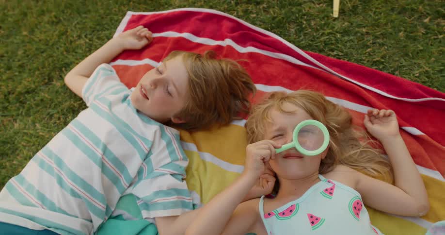 Two children lying on a red and white striped blanket on grass their heads close together
