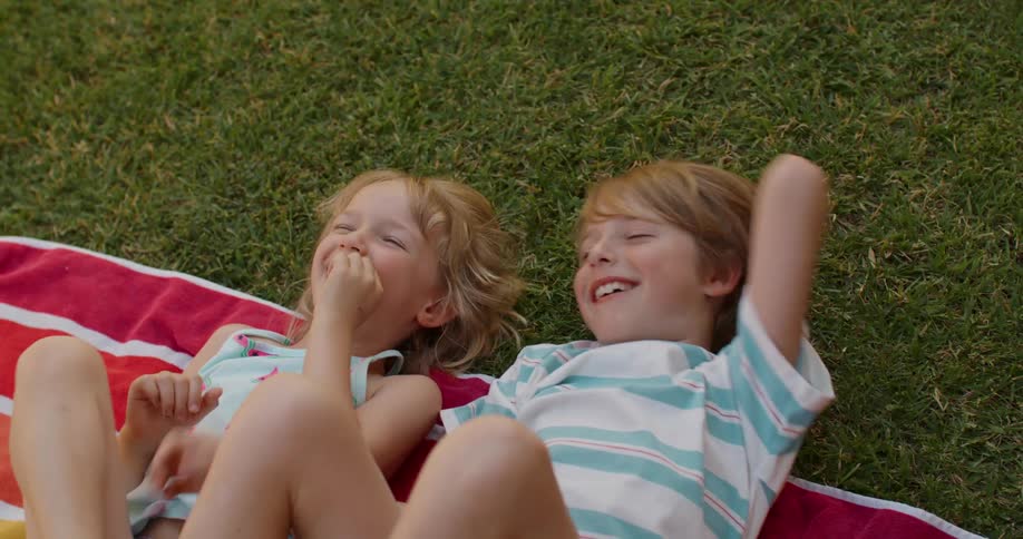 Brother and sister lie on a towel on the grass and laugh together
