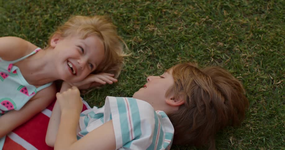 Brother and sister lie on a towel in the grass and talk as they point around