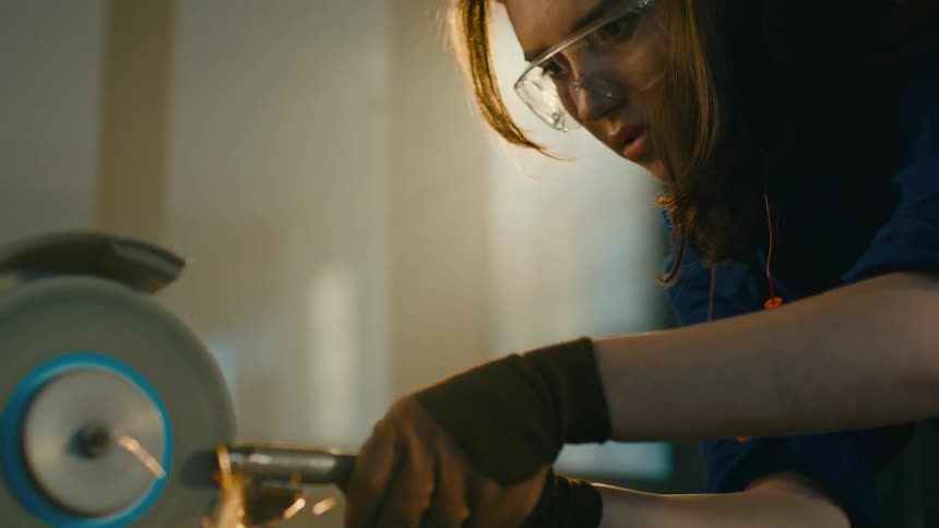 An woman wearing gloves is grinding a piece of metal with a power tool in a workshop