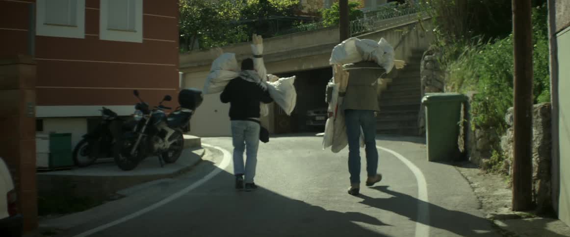 Two men carry bundles of sticks down a road