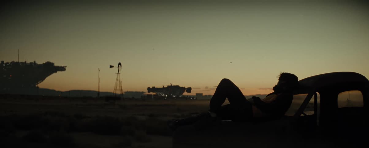 Person sits on their car and leans against the rear window as the sun sets near a space port
