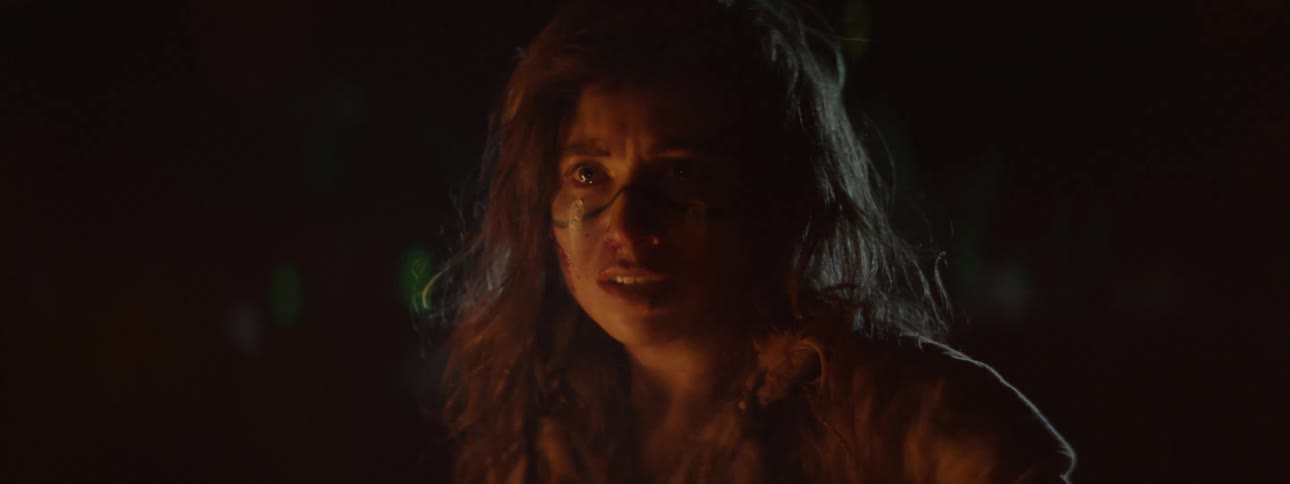 A young woman sits at a campfire and tears up while talking