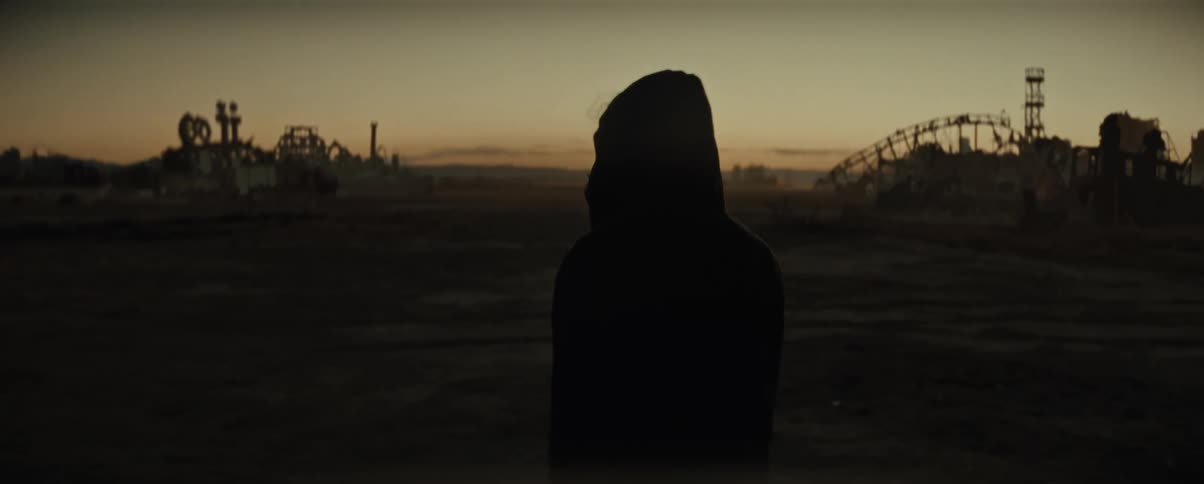 A male with a hoodie walks across an open field near broken down structures