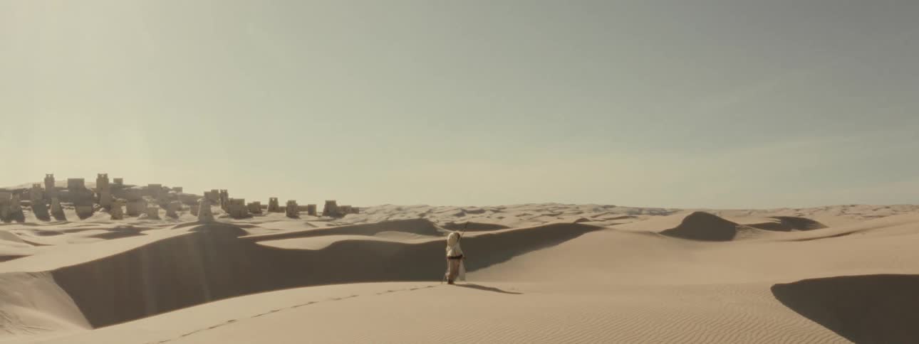 Woman walks through the desert with a field of towering structures in the background