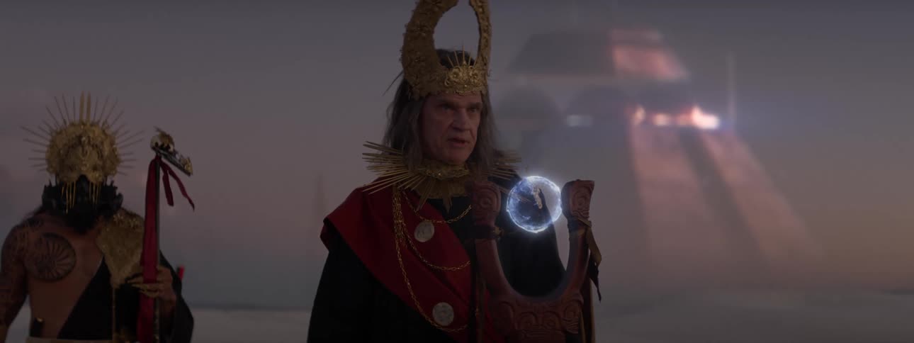 A person is wearing golden clothes and speaking and in front of him is a staff with a glowing blue orb on top