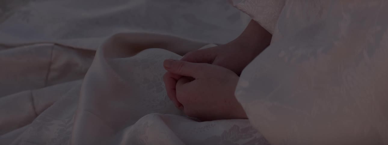 A closeup of clasped hands covered by white lacy fabric resting on a lap