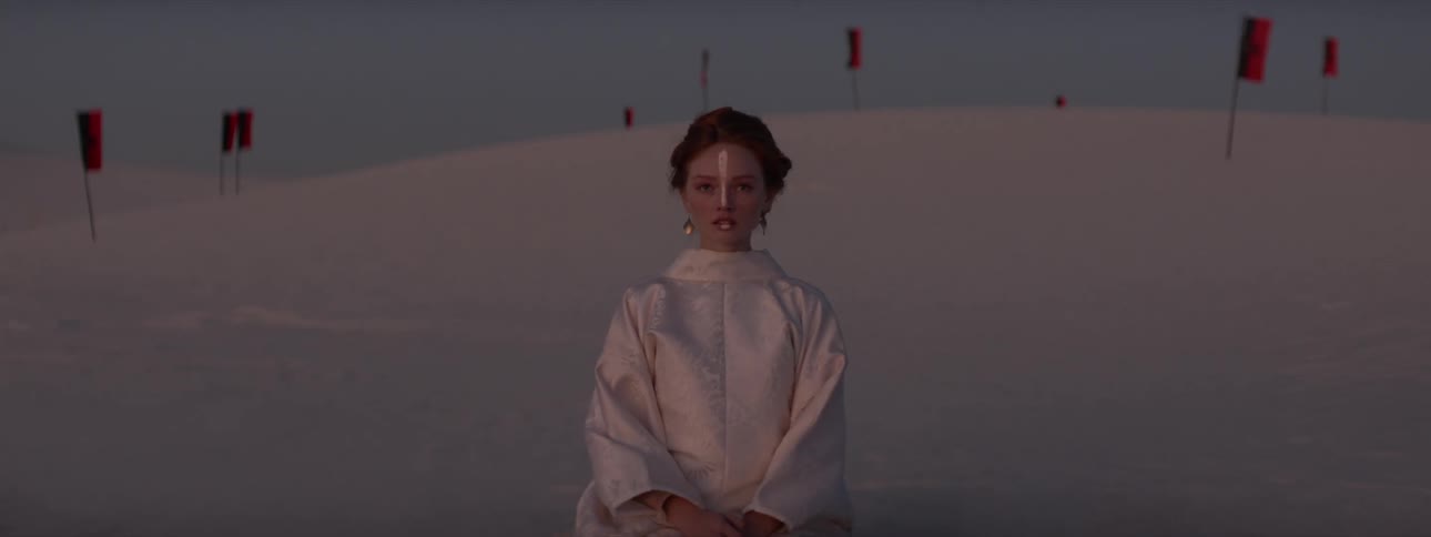 Woman kneeling in white kimono in desert dunes with flags in the background