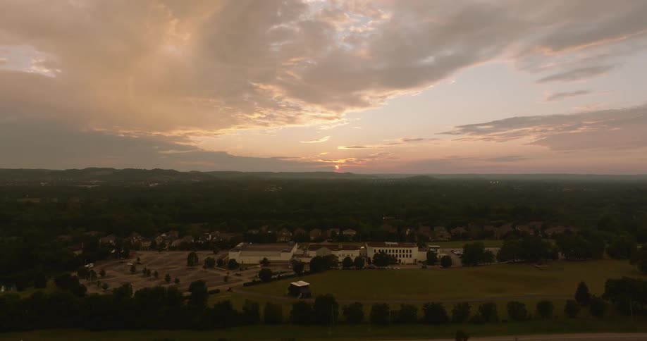 Aerial panorama of buildings surrounded by a vast field with trees against a sunset sky