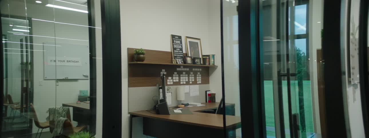 Modern office environment with glass walls and doors highlighting an interior design that includes a wooden bookshelf with folders a desk with a computer and plants