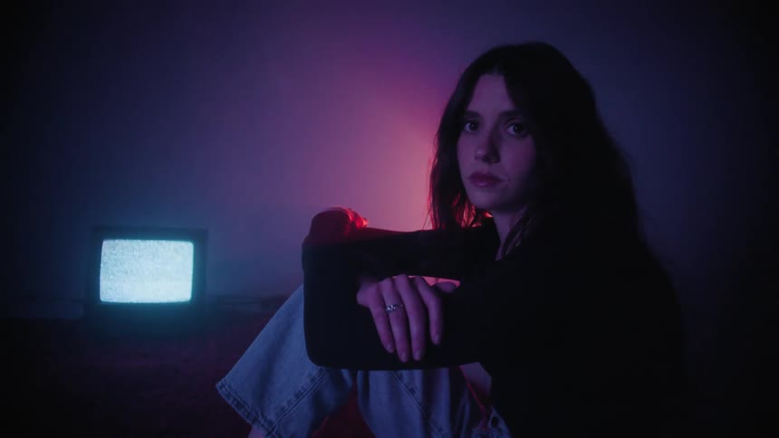 A young woman sitting on the bed in a dark room with a red light coming from behind her