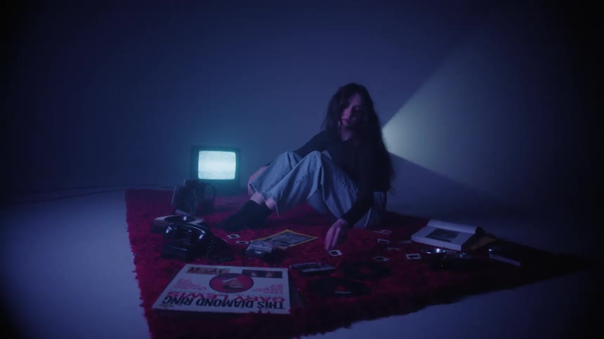 A young woman sitting on a red carpet surrounded by vinyl records video tapes and cameras on a dimly lit room