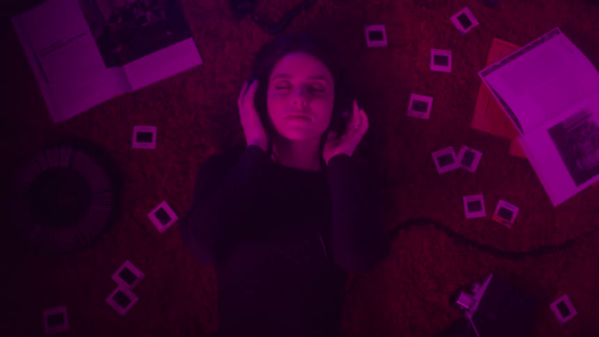 A young woman listening to music on headphones and she is lying on a red carpet with several photos on her sides