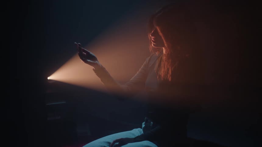 A young beautiful woman sitting in a dimly lit room with a beam of light shining onto their hand from the left side