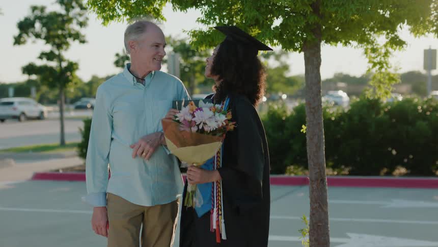 Father next to his daughter who is wearing a graduation gown and holding a bouquet of flowers smiling