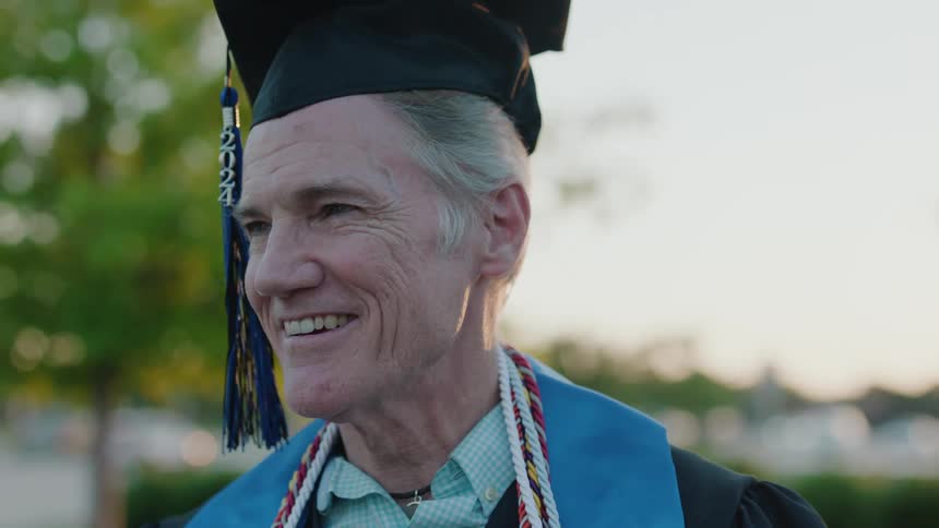 A mature man wearing a graduation cap and gown adorned with a colorful cord