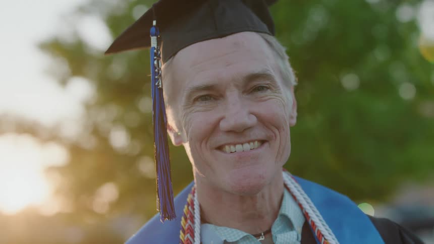 An elderly man wearing a graduation cap is smiling widely
