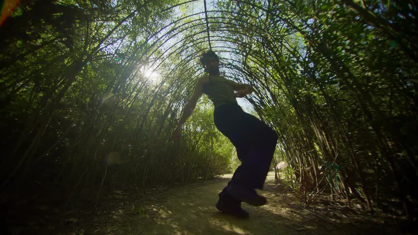A man performs an exaggerated dance while standing in a tunnel made out of young trees