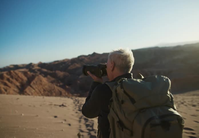 Man walking in a desert terrain with dunes in the background during the day and using a professional camera