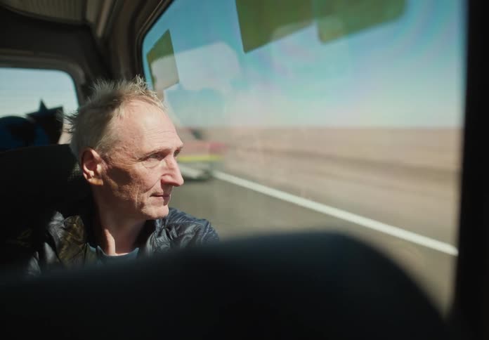 An elderly man seated inside a bus gazing out the window to a desert landscape