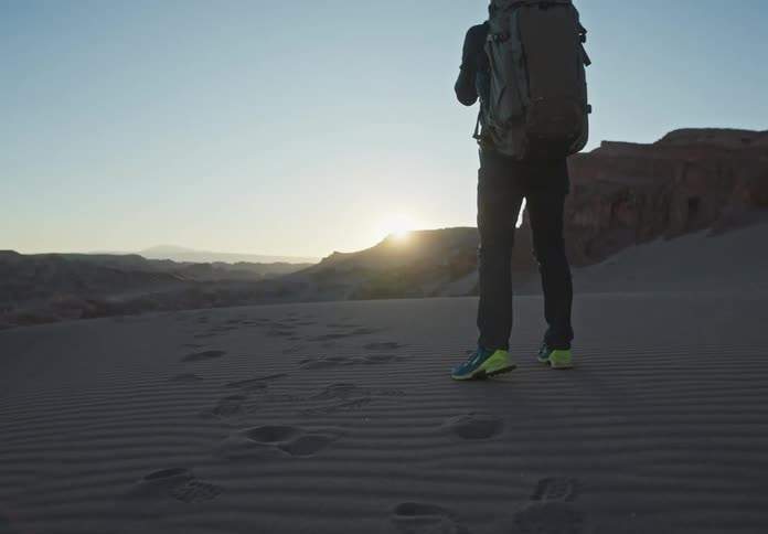 Male backpacker walks back and forth on sand while taking pictures of the sun on the horizon