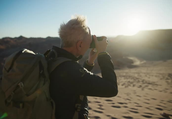 A grayhaired man with a backpack is taking photos in a desert region with strong sunlight coming from above