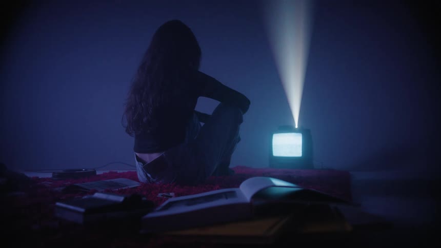 A woman sitting on a bed looking at a tv with a white screen and a red light playing over her