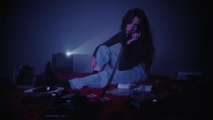 A young woman sits on the floor in a dimly lit room holding a vintage phone
