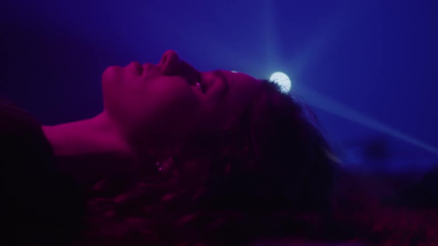 Side portrait of a woman lying on a red carpet in a dark room with a neon light in the background