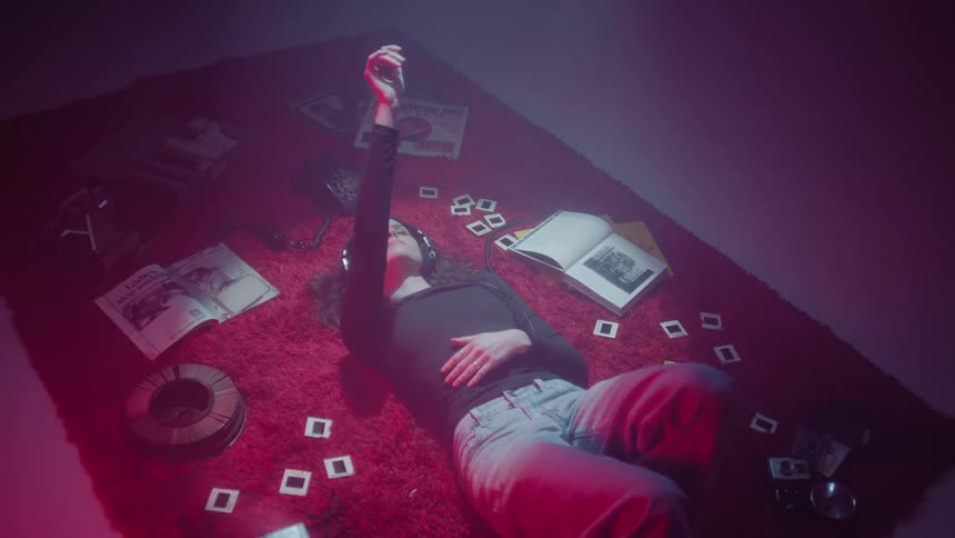 A young woman lying on a red carpet with several photos next to her and she is wearing headphones and moving