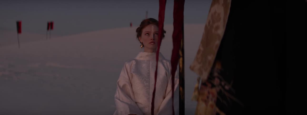 A woman wearing a shiny robe sits on the desert surrounded by flags looking up at a robed figure