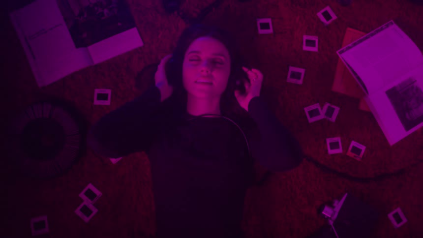 A young woman lying on a red carpet with several small photos spread out behind her and she has headphones on and listening to music