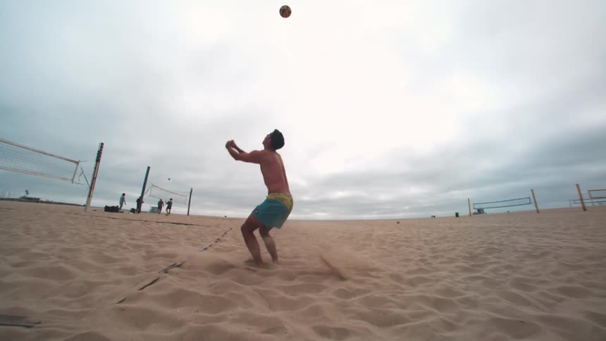 A shirtless man is throwing a ball in the air and hitting it with his hand on the sand of a beach volleyball court
