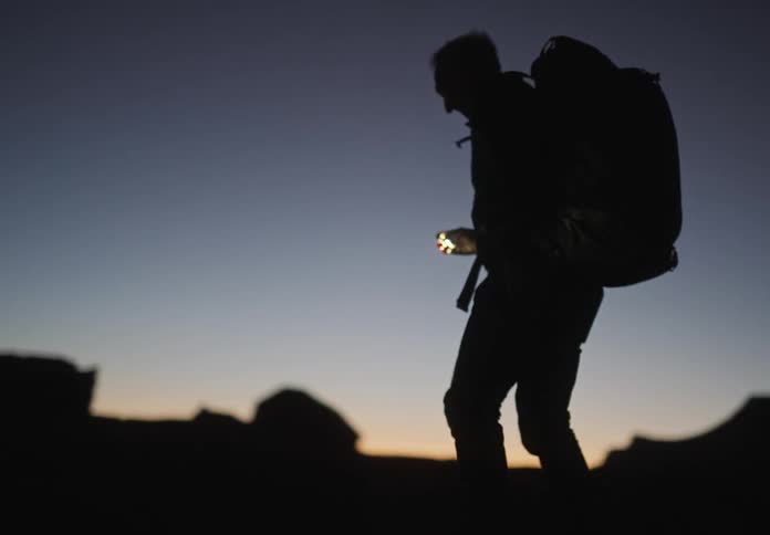 Silhouette of a man with a backpack holding a flashlight and a professional camera walking through the mountains at dawn