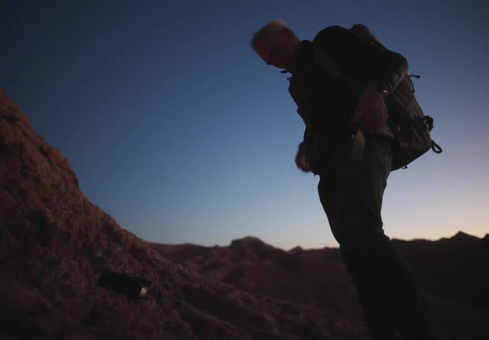 Man putting backpack on his back while standing in a natural environment with rocks and mountains around