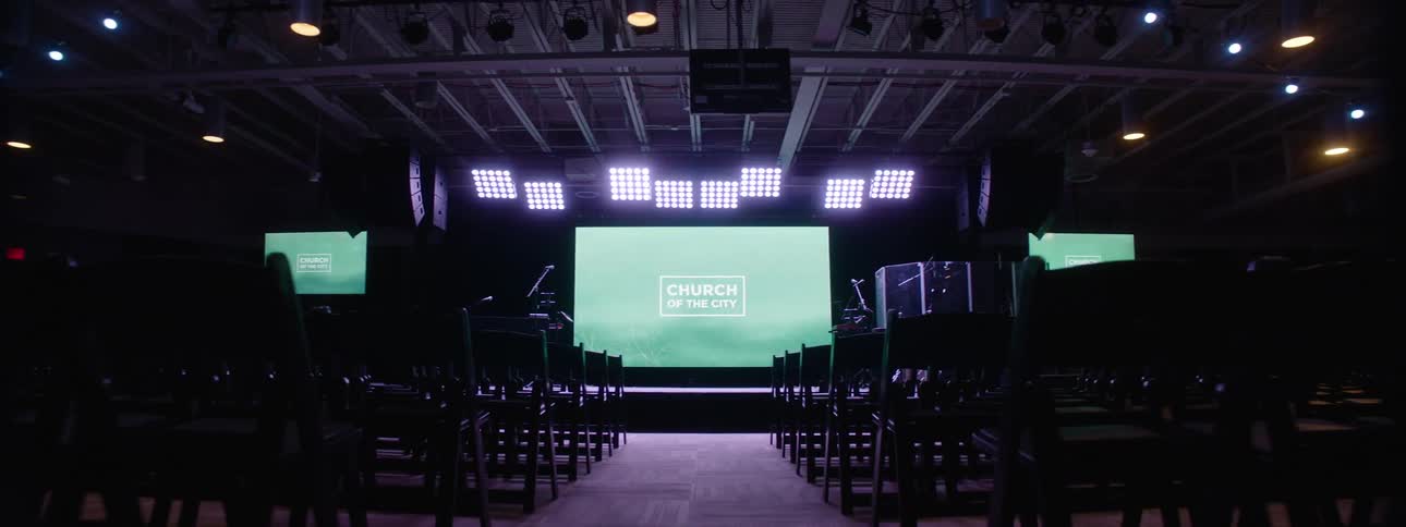 Dark interior with rows of black chairs facing a stage lit by overhead lights and projectors
