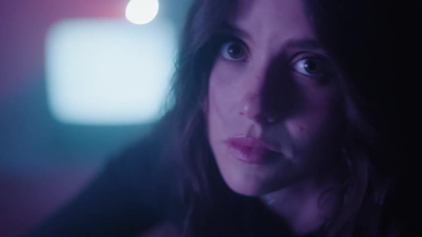 A dark haired woman looks fearful as a tv and flashing light play in the background