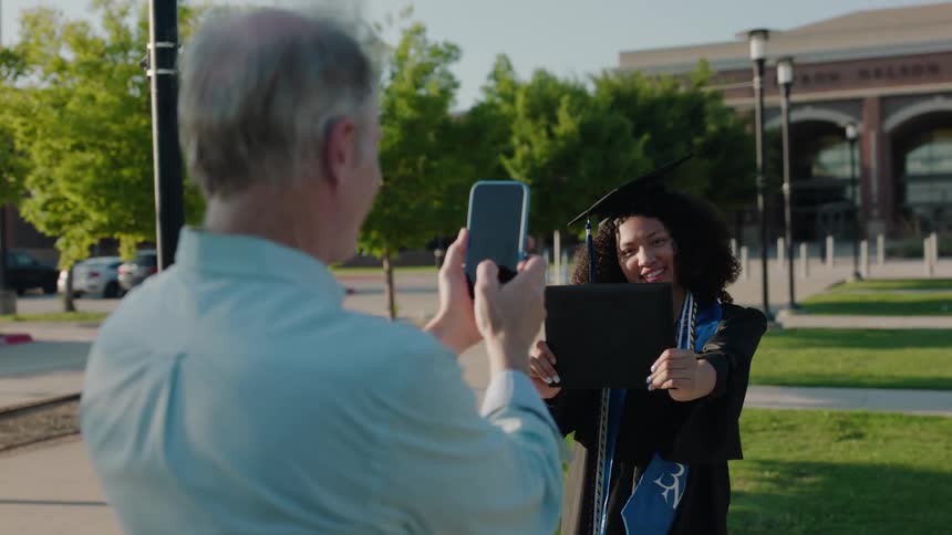 Father snapping a picture of daughter holding out her diploma from cell phone