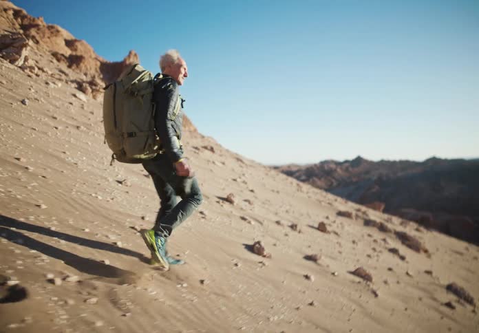 Man walking on a desert dune with mountains around carrying a large backpack on his back and holding a camera